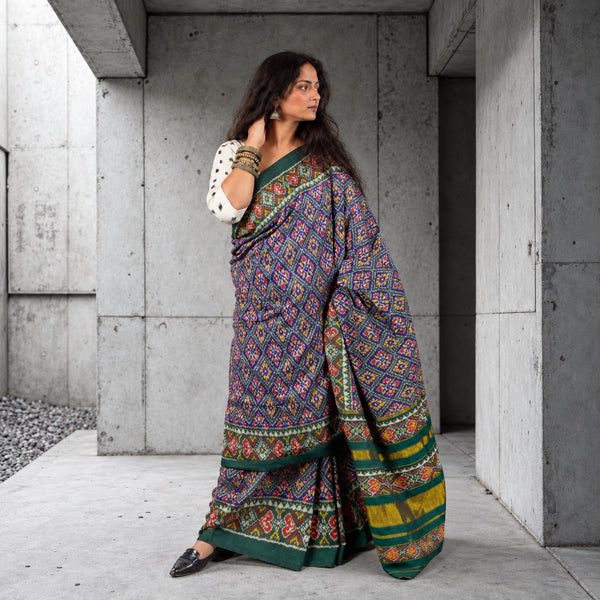 A woman wearing a dark purple and green patterned saree with a white polka-dot blouse, standing in a concrete corridor.